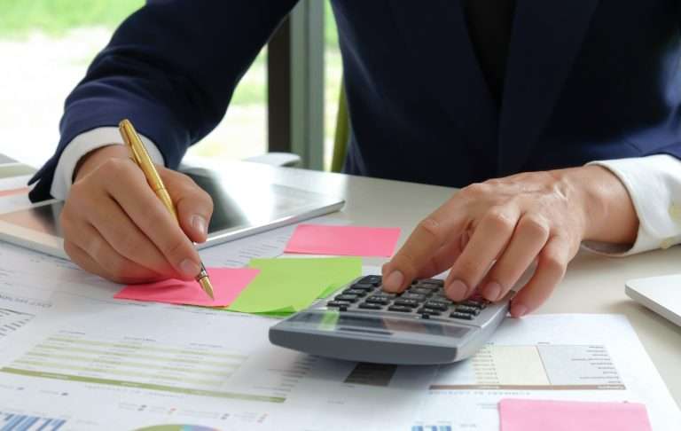 Um profissional em traje formal avalia documentos financeiros para a análise de propostas sob a nova lei de licitações, utilizando uma calculadora e fazendo anotações em notas adesivas coloridas, entre gráficos impressos e gráficos.