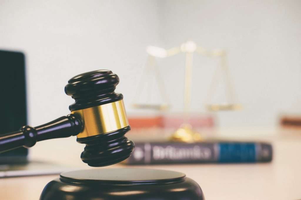 Martelo de juiz em destaque sobre superfície escura com balança de justiça ao fundo e livro intitulado “Diccionario”, simbolizando o princípio da isonomia nas licitações.