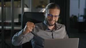 Pessoa de camisa social cinza sentada à mesa em frente ao laptop em ambiente de escritório escuro, fazendo gesto enfático com o punho, representando possivelmente uma vitória na licitação na modalidade pregão.