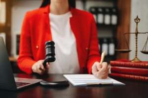 Profissional jurídico em blazer vermelho trabalhando em uma mesa com martelo, caneta, laptop, livros de direito e balança da justiça, simbolizando autoridade regulatória e mecanismos de controle na nova legislação
