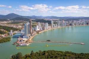 Vista aérea de Balneário Camboriú, em Santa Catarina, mostrando a orla da Praia Central com seus altos edifícios, um píer avançando sobre o mar e o rio Camboriú com uma ponte estaiada, evidenciando o desenvolvimento da região e as oportunidades de licitações em Santa Catarina.