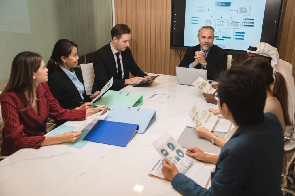 Sete pessoas sentadas ao redor de uma mesa de reunião, analisando documentos com gráficos. Um consultor de licitação lidera a conversa, com tela de apresentação ao fundo.