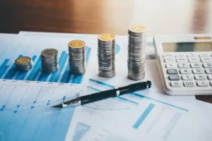 Pilhas de moedas organizadas em escala crescente sobre relatórios com gráficos financeiros e calculadora ao lado, representando a análise de indicadores fiscais e capacidade de pagamento (CAPAG) de estados e municípios antes da celebração de contratos públicos.
